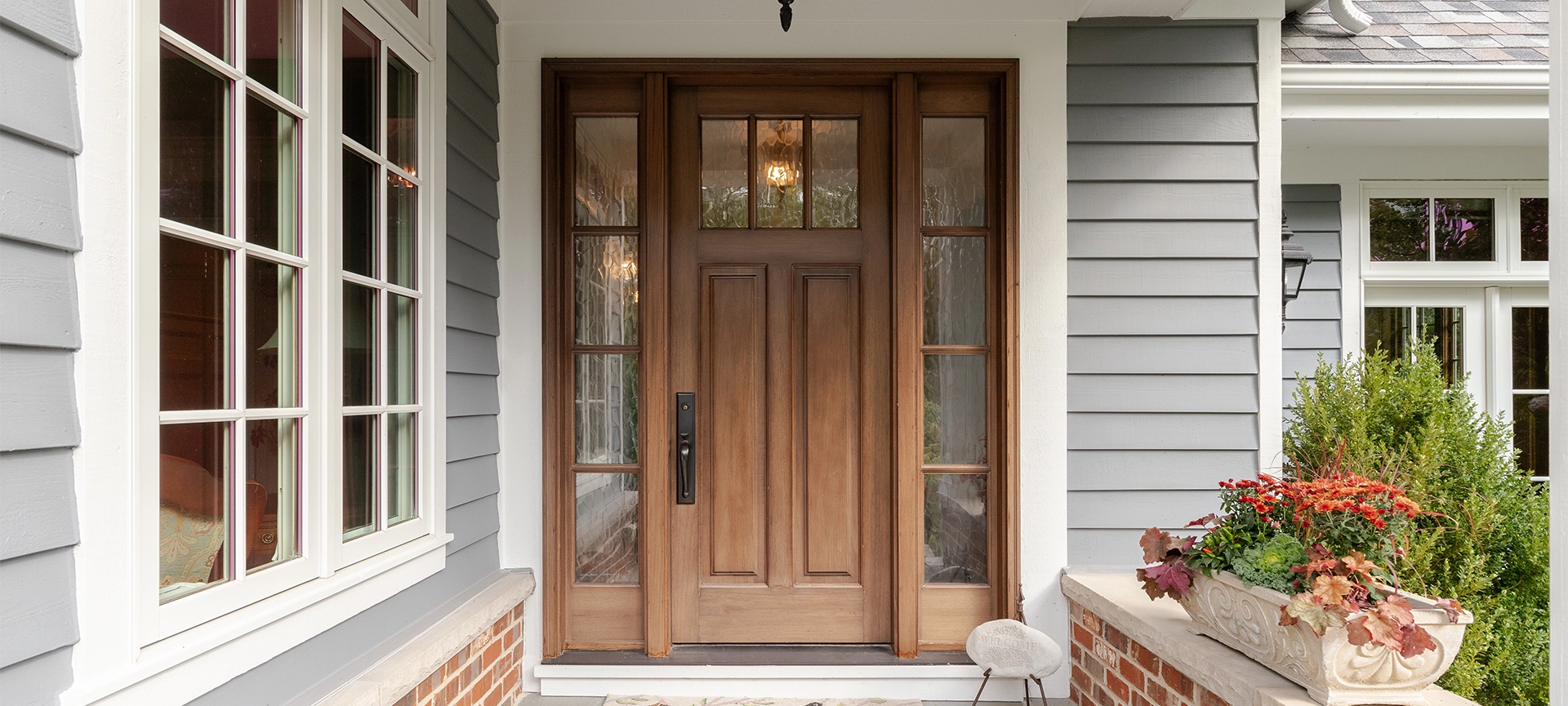 wooden front door and covered porch detail on a home with grey siding, white trim, red brick