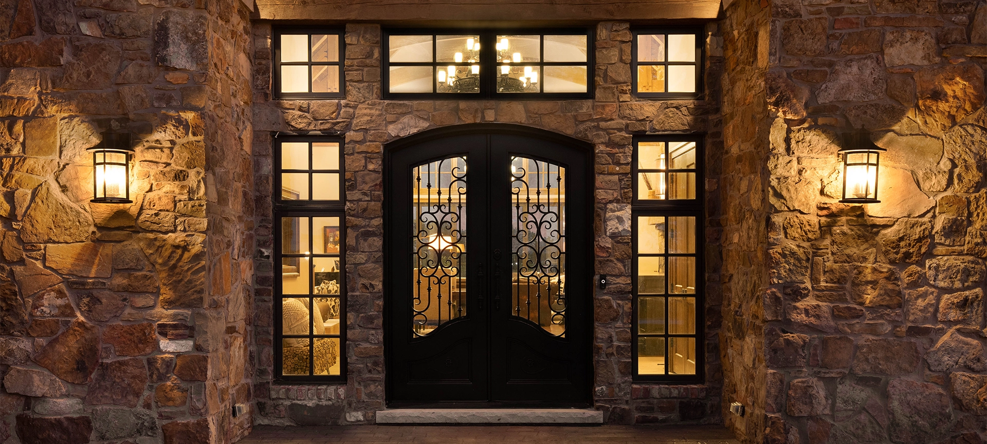 A luxury home with a fancy iron door, surrounded by windows and a rock exterior. Warm bulbs light the outside and inside of the property at sunset.