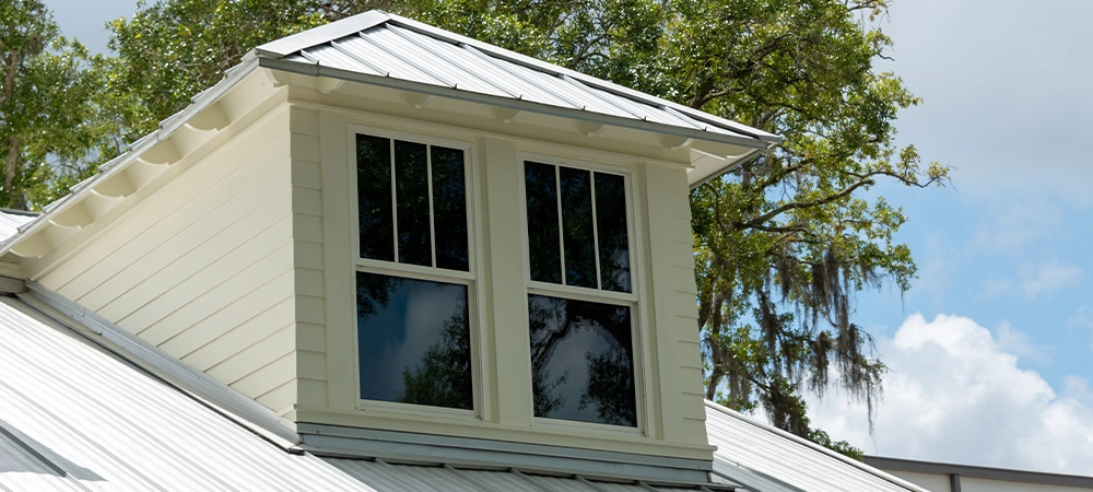 A dormer with multiple double hung windows, a grey metal roof, and pale yellow vinyl siding on a large house. There are tall trees with thick green leaves both in front and behind the building.