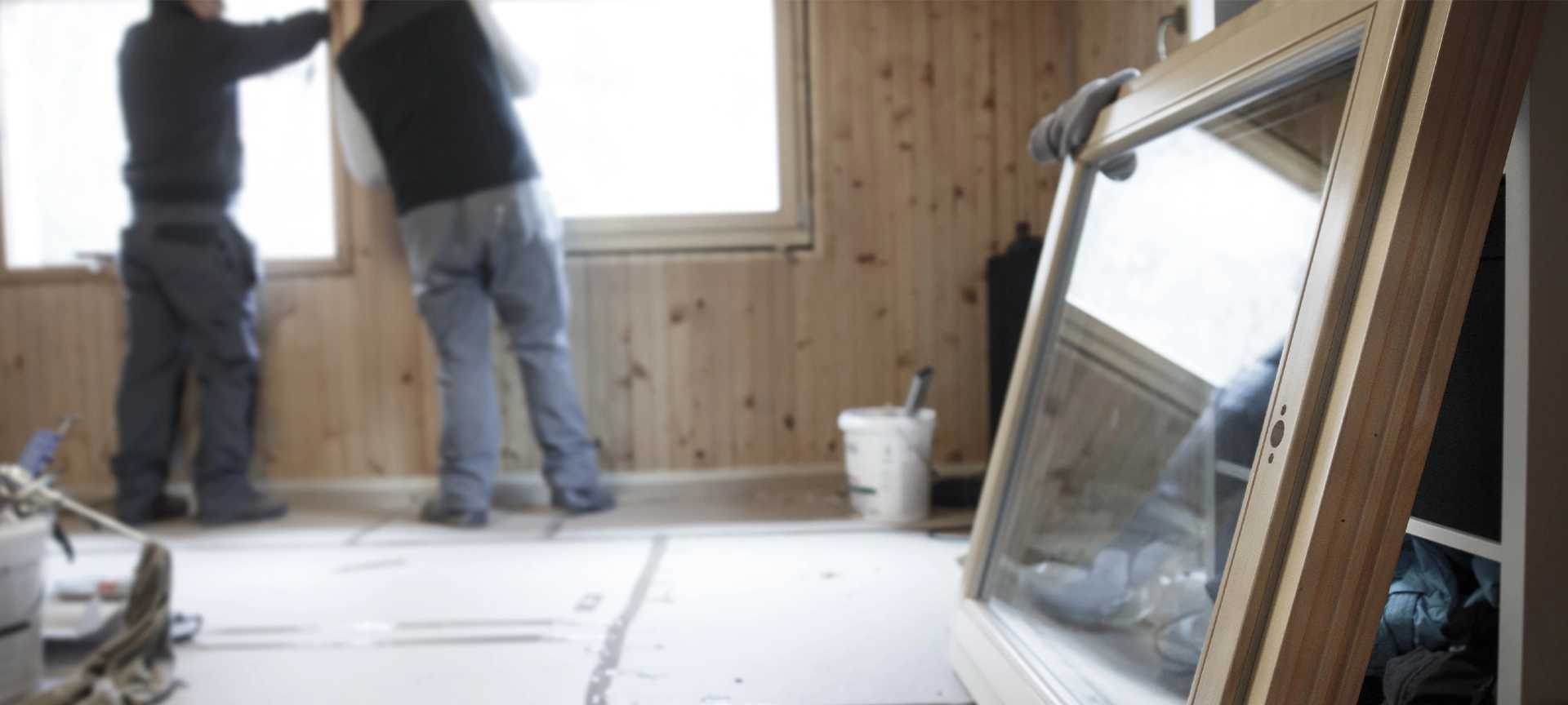 Workers in the background installing new, three pane wooden windows in an old wooden house, with a new window in the foreground. Home renovation, sustainable living, energy efficiency concept.