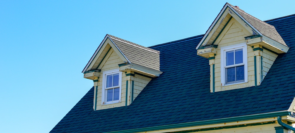 The top exterior floor of a yellow colored Cape Cod house with green and white trim. The building has two peaked dormer windows. The roof is covered in black asphalt shingles. The sky is bright blue.