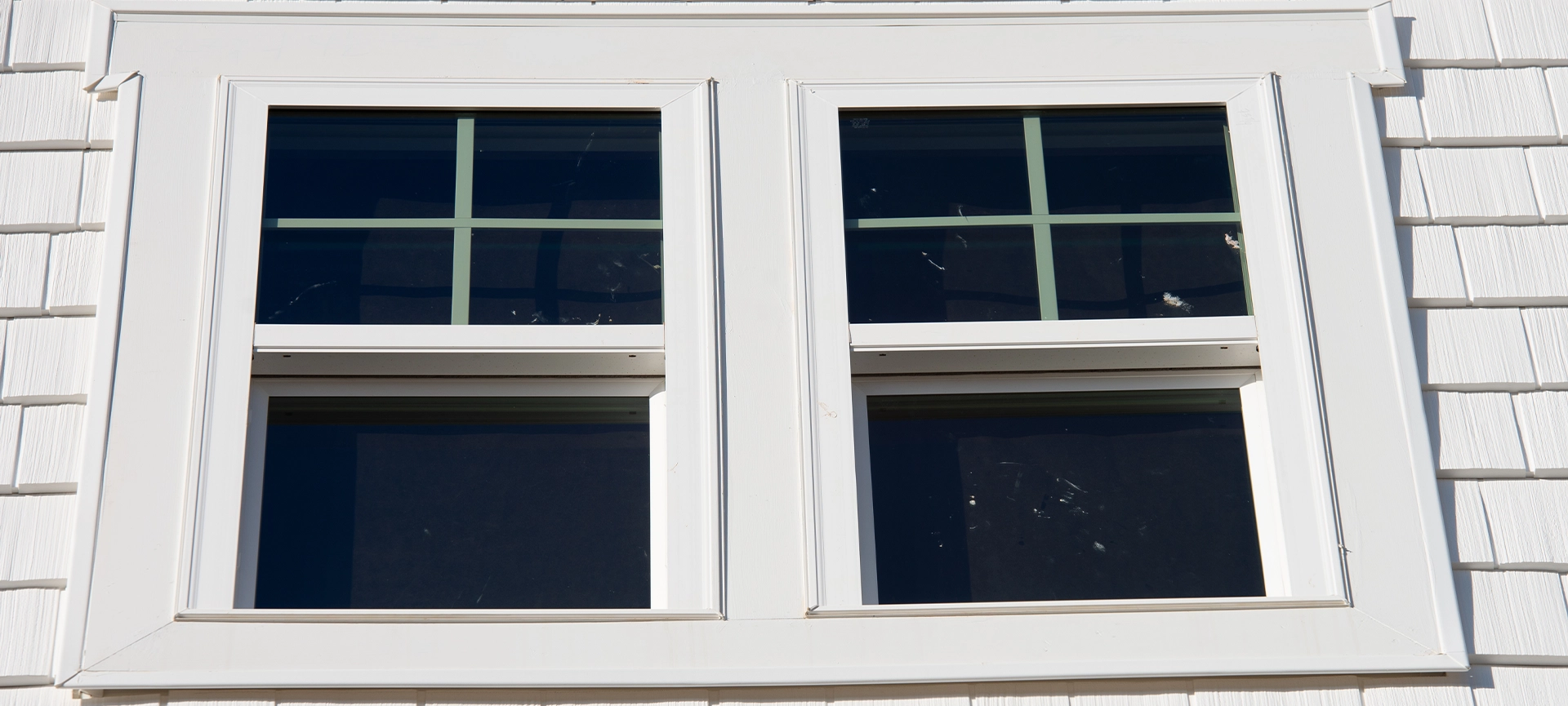 Double hung window with fixed top sash and bottom sash that slides up, sash divided by white grilles a surrounded by white elegant frame horizontal white shingle siding on new construction residence